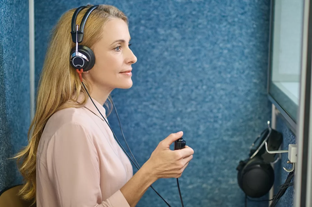 An older women sitting in soundproof booth at a hearing clinic wearing headphones and taking a air conduction threshold hearing test